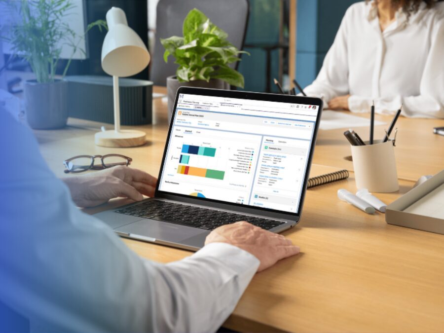 Person working at a desk on a laptop displaying bar charts and data dashboards, engaged in scientific publications planning, with office supplies and another person in the background.