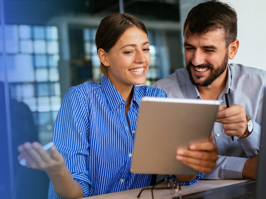 Two people sit together looking at a tablet, smiling and discussing something. One person gestures with their hand while the other holds a pen.